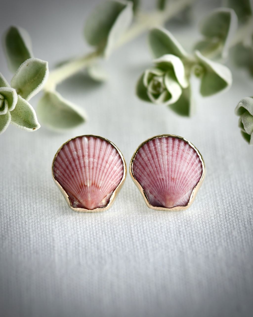 Pink scallop shell earrings on a white fabric background with greenery