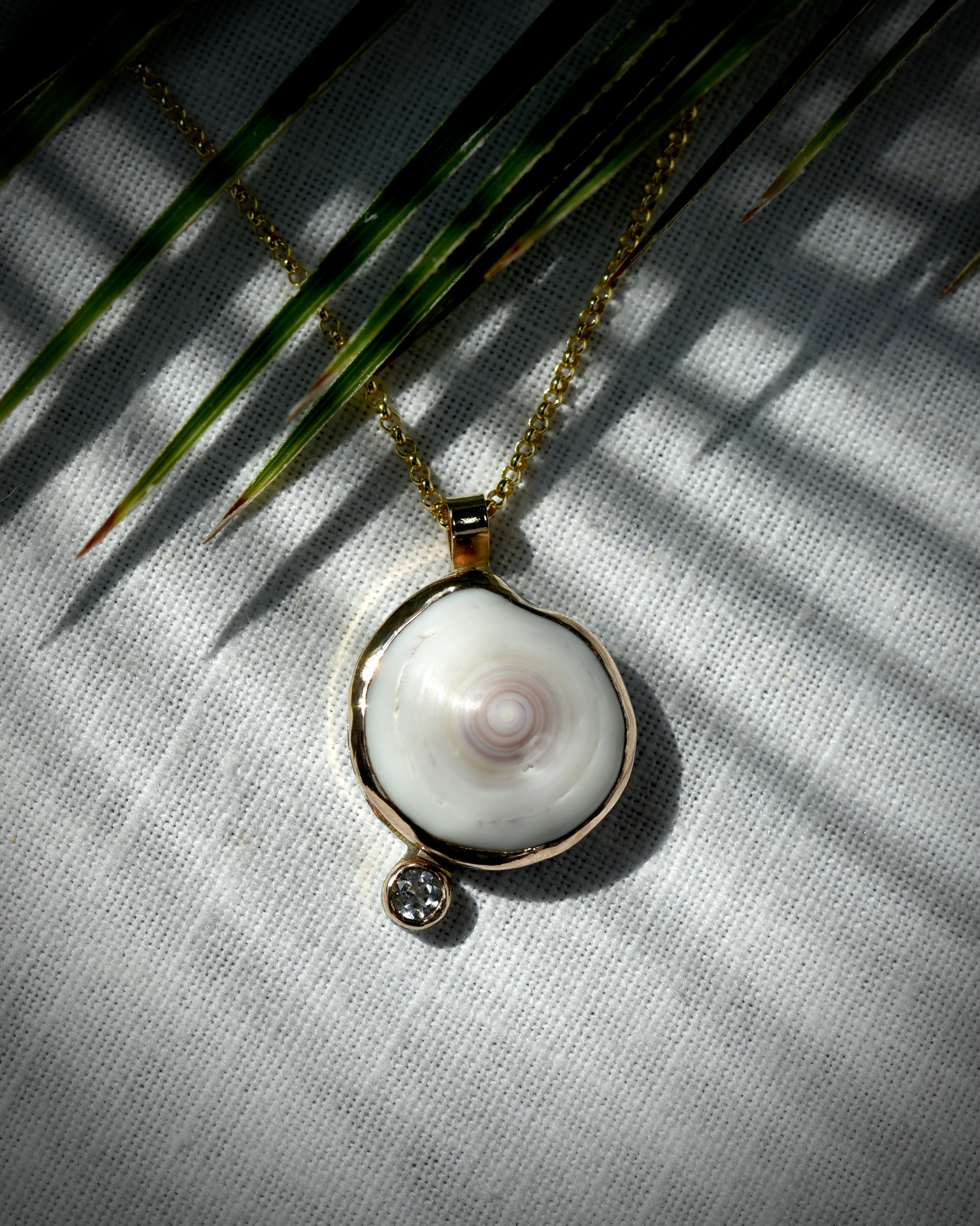 Close view of luminous white Hawaiian puka shell and topaz Midi Puka Dot necklace displayed beneath palm leaves