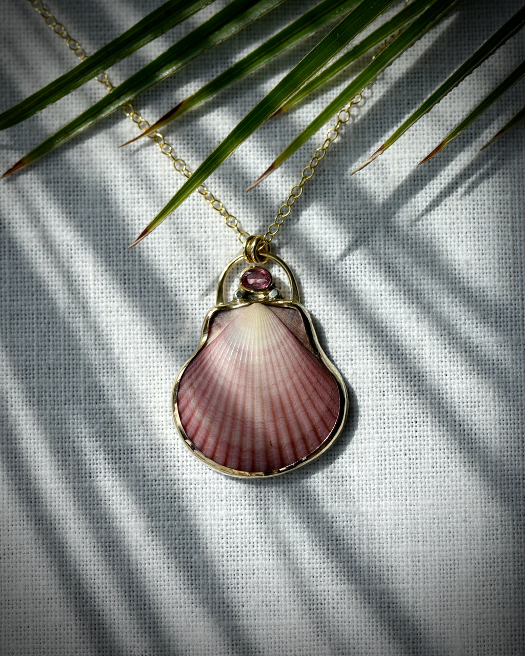 Close view of pink scallop shell pendant necklace displayed beneath palm leaves