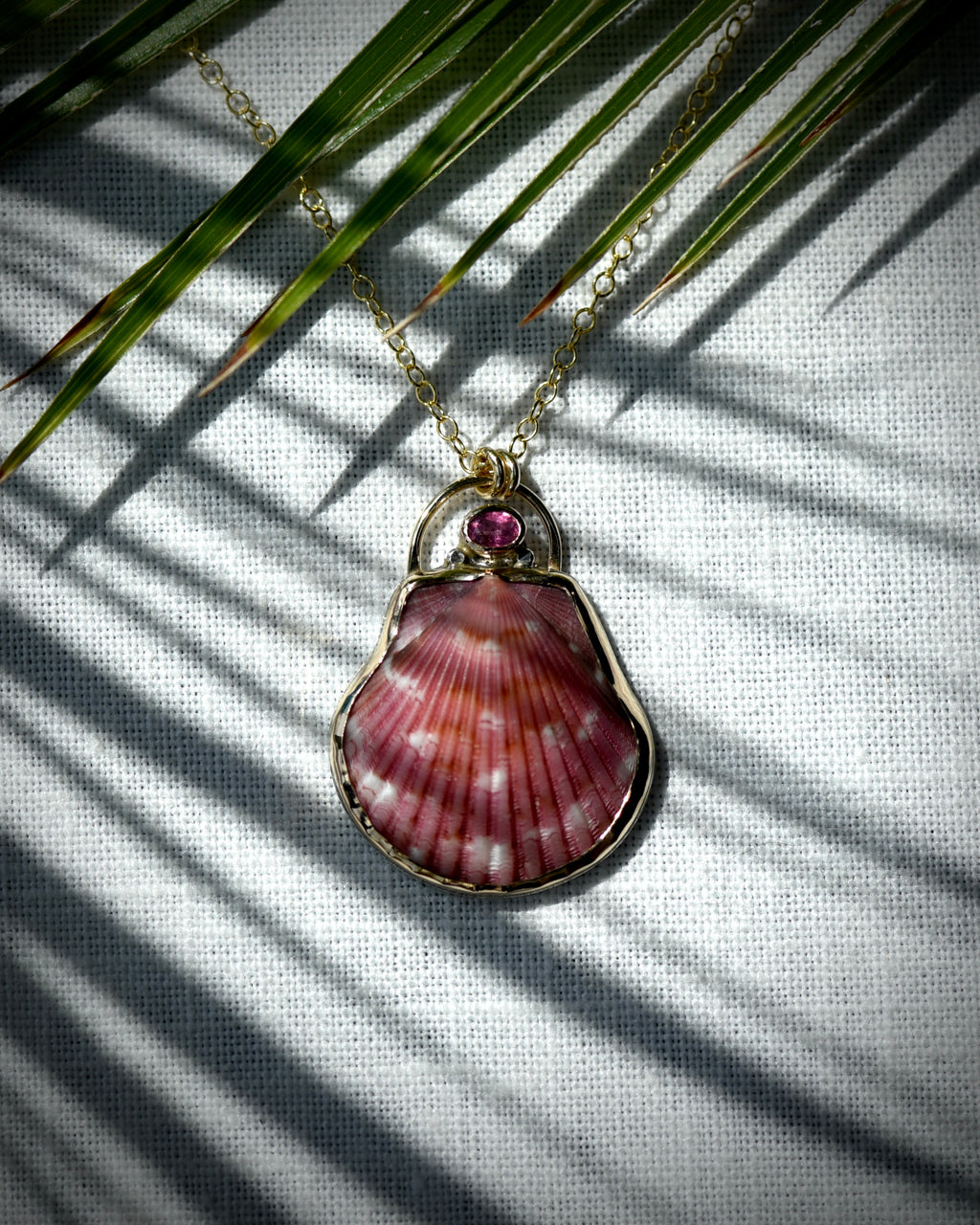 Close view of deep pink scallop shell pendant necklace displayed beneath palm leaves
