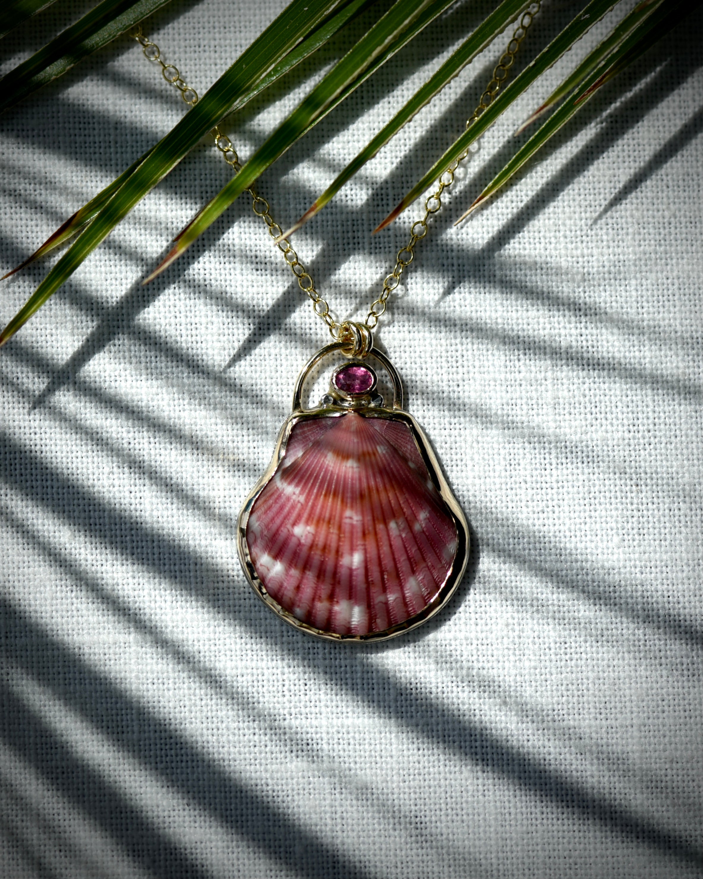 Close view of deep pink scallop shell pendant necklace displayed beneath palm leaves

