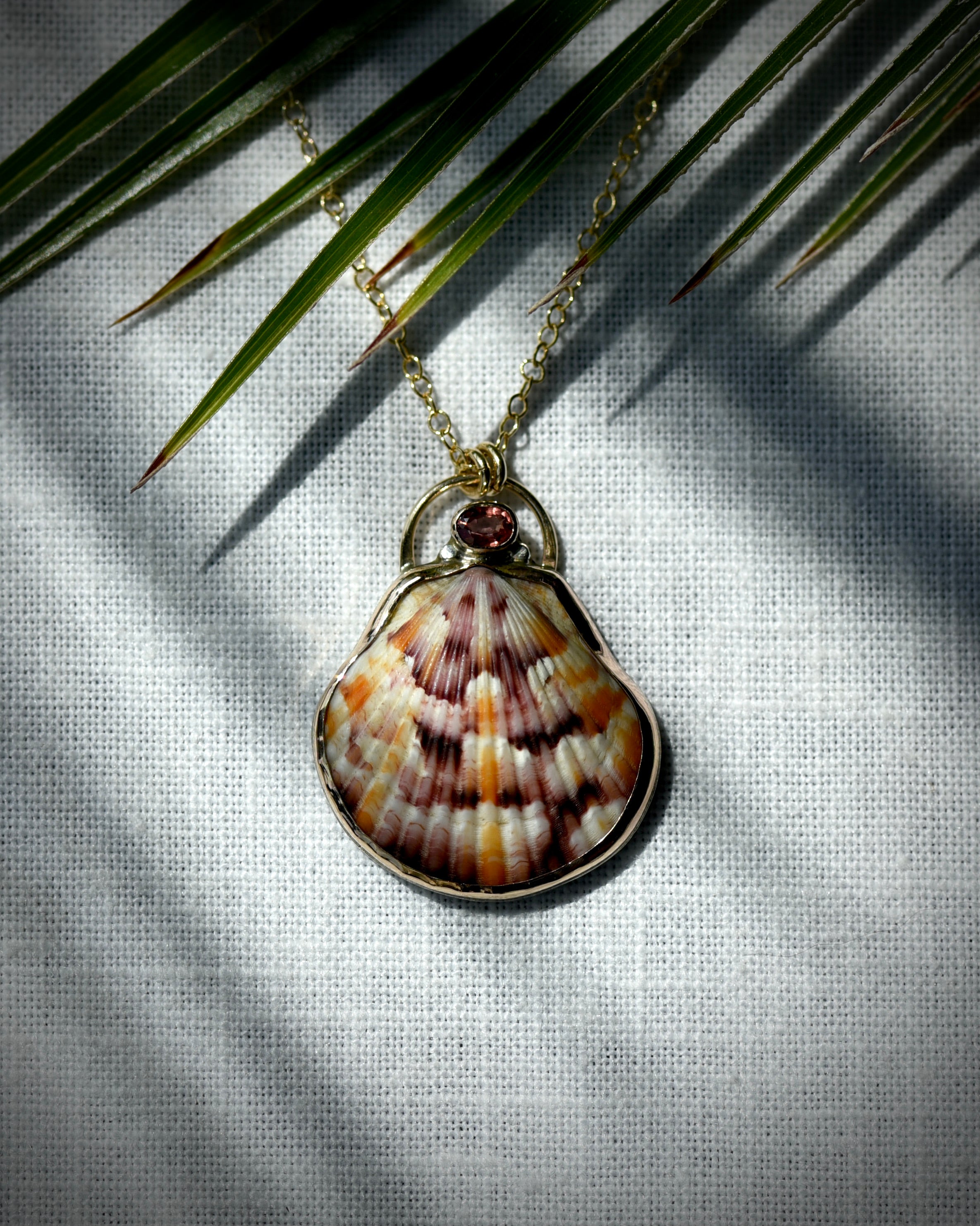 Close view of patterned scallop shell pendant necklace displayed beneath palm leaves